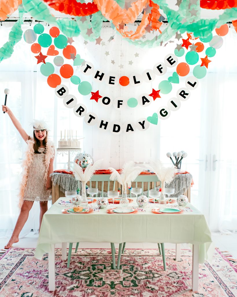 Showgirl birthday party decor inspired by Taylor Swift’s Life of a Showgirl album. Tween girl dressed in sparkly showgirl outfit and feather boa stands beside a mint and orange party table with disco balls, feathers, and a banner reading ‘The Life of a Birthday Girl’
