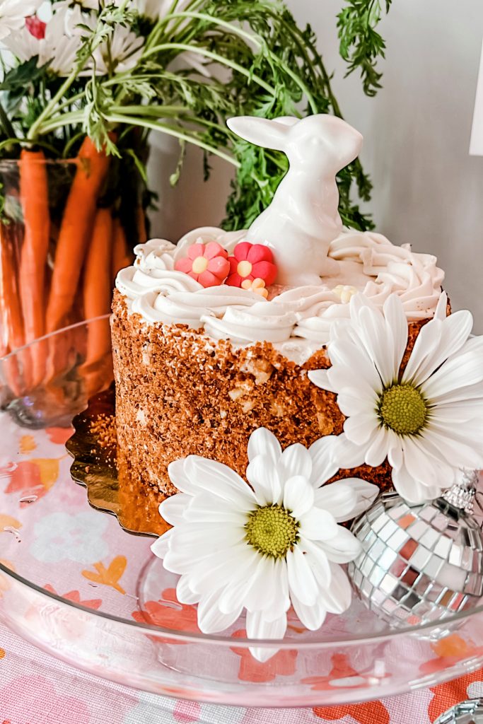 A whimsical bunny birthday cake made by decorating a store-bought carrot cake with a ceramic figure, flower sprinkles, disco ball, and daisies. 