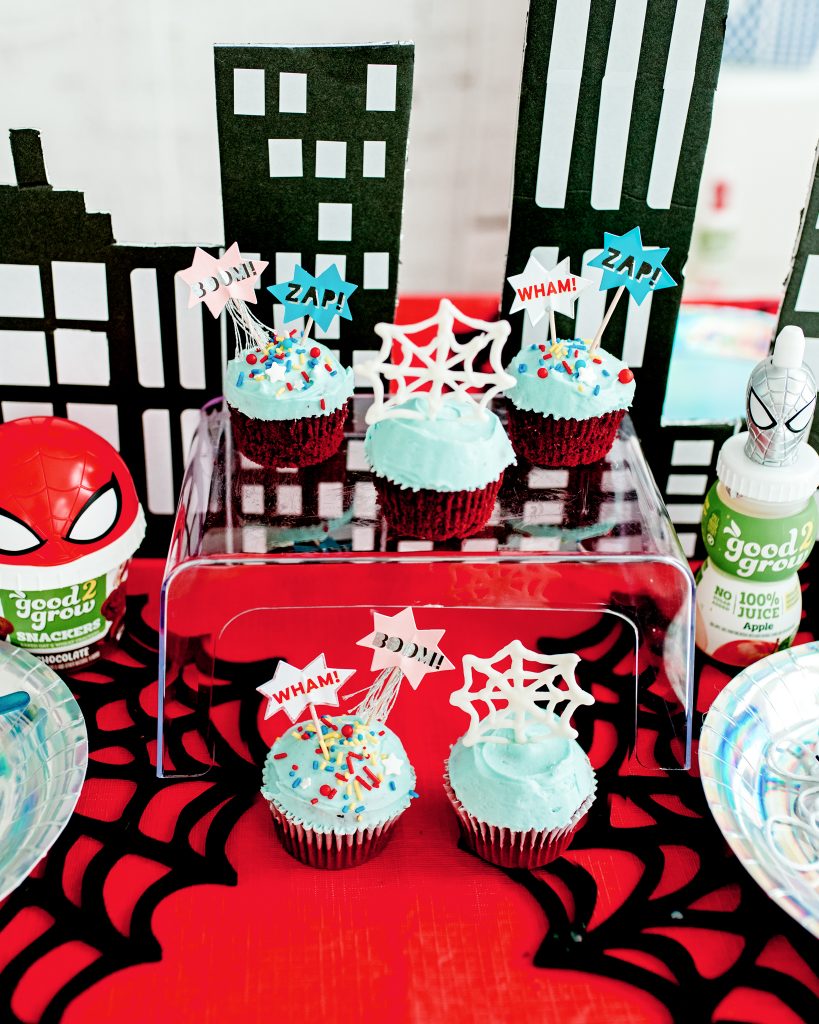 Red velvet cupcakes with blue frosting topped with chocolate spiderwebs and superhero toppers on a dessert tray. DIY chocolate spiderweb toppers